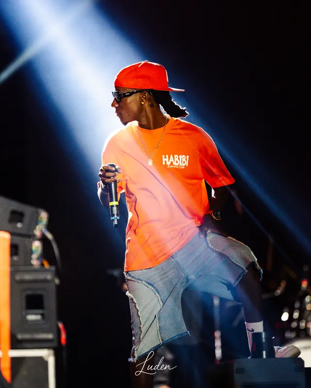 Un chanteur casquette rouge et t-shirt orange au micro, traversé par un faisceau de lumière.