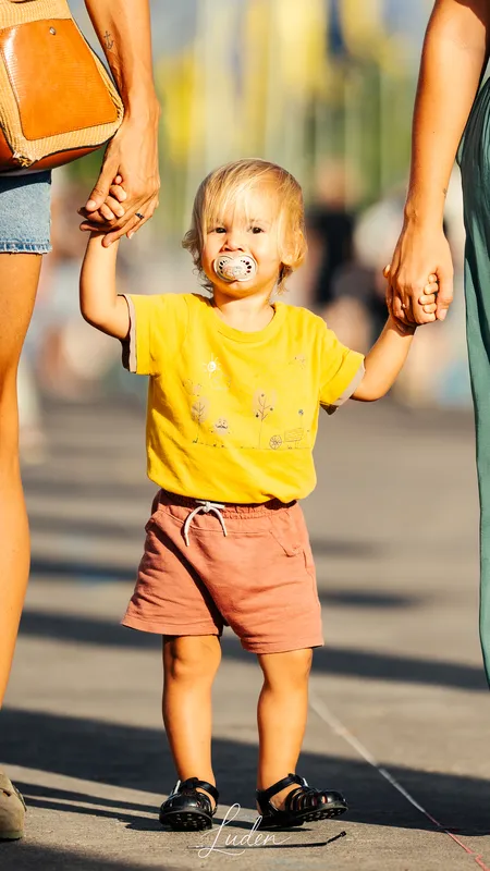 Un jeune enfant en t-shirt jaune marche dans la rue, tenu par la main par deux adultes.