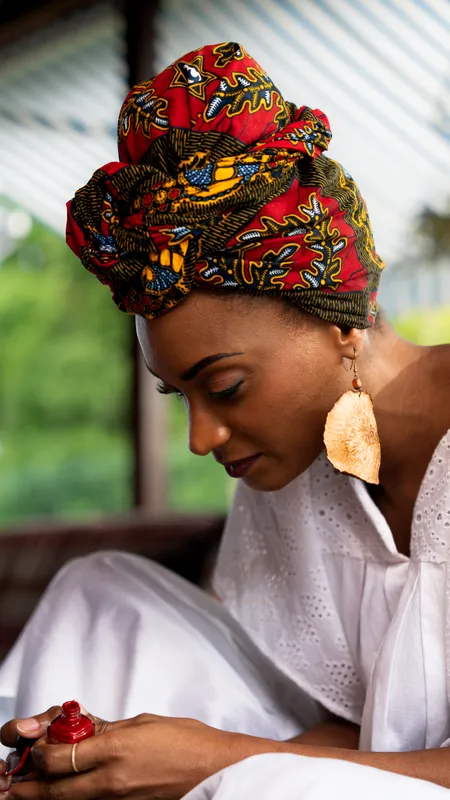 Une femme coiffée d’un foulard wax rouge et jaune, le regard baissé, de grandes boucles d’oreilles dorées.