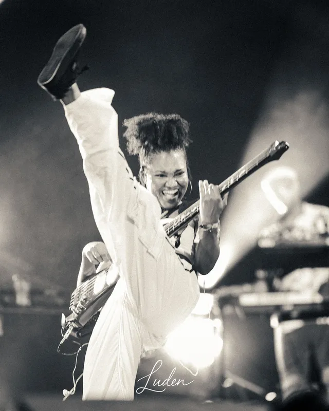 Un guitariste tout en blanc, la jambe levée, en plein élan sur scène. Noir et blanc.