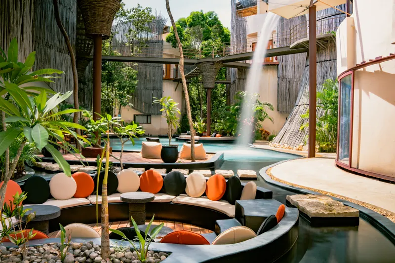 Un salon extérieur aux coussins colorés, au bord de l’eau, avec une cascade.